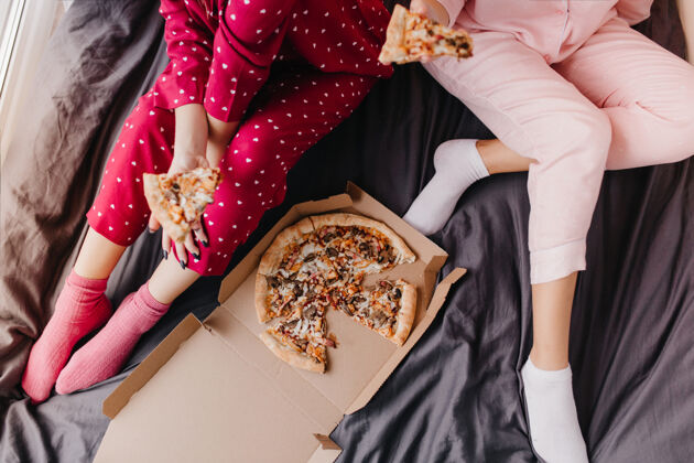 饥饿两个穿着睡衣的女孩坐在床上吃意大利快餐的头像懒惰的女模特在深色床单上吃比萨饼头顶朋友欢乐