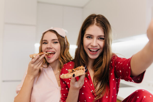 用餐情绪化的卷发女孩微笑着吃着比萨饼室内照片是身着红色睡衣的快乐棕色头发女人和朋友自拍的照片朋友睡衣休闲