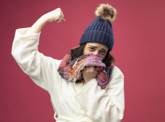 人虚弱的生病的年轻女子戴着长袍 戴着冬天的帽子和围巾 用围巾捂住嘴 看着前面做着强烈的手势 孤立在粉红色的墙上衣服姿势公民