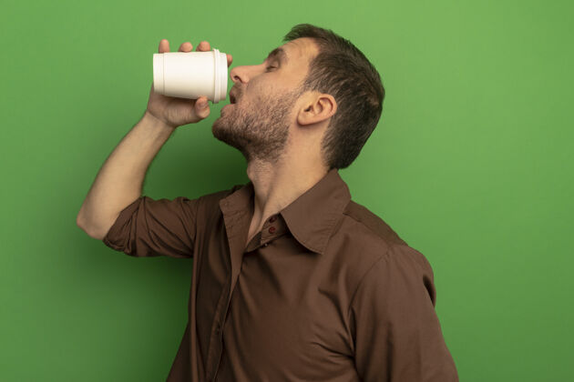 表情年轻人站在侧视图喝咖啡从塑料咖啡杯隔离在绿色墙上姿势喝酒轮廓