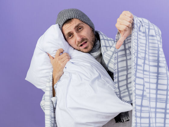 格子悲伤的生病的年轻人戴着冬天的帽子 围巾裹在格子布的抱枕里 在紫色的背景上孤立地竖起大拇指围巾拇指帽子