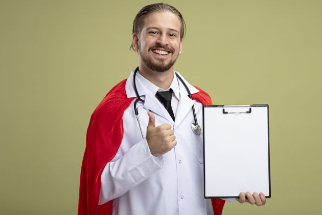 剪贴板年轻的超级英雄微笑着 戴着听诊器 穿着医用长袍 拿着剪贴板 在橄榄绿的背景上孤立地竖起大拇指微笑医疗抱着