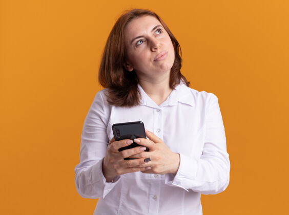年轻身穿白衬衫的年轻女子手持智能手机 带着沉思的表情 站在橙色的墙上 思考着积极的一面抱着在一旁衬衫