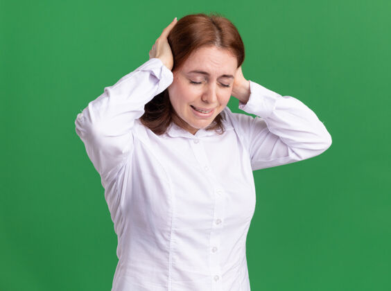 触摸一位身穿白衬衫的年轻女子站在绿色的墙壁上摸着头痛的头 看上去很不舒服年轻头痛女人