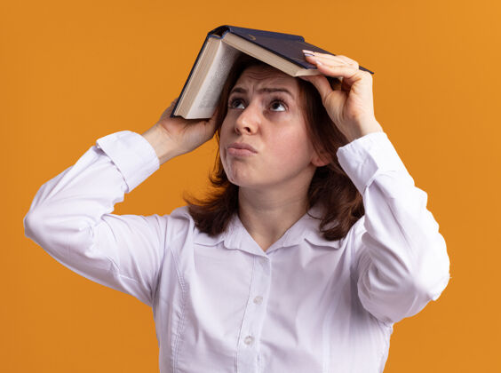 年轻身穿白衬衫的年轻女子头上举着一本打开的书 站在橙色的墙上困惑地抬头看女人困惑头