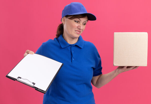 女人身穿蓝色制服 头戴帽子的中年女送货员站在粉红色的墙上 手里拿着纸板箱和剪贴板 空白的书页看上去既困惑又不高兴交货制服混乱