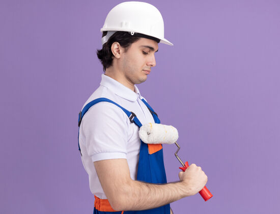 建筑年轻的建筑工人穿着建筑制服 戴着安全帽 手里拿着油漆辊 站在紫色的墙上建筑工人站着制服
