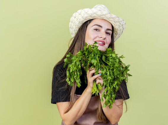 请高兴美丽的园丁女孩穿着制服 戴着园艺帽 手持香菜 脸上孤立的橄榄绿背景帽子橄榄花园