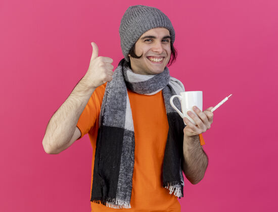 抱着带着微笑的年轻病人戴着冬天的帽子 戴着围巾 手里拿着一杯茶 温度计在粉色背景上孤立地竖起大拇指微笑年轻人围巾