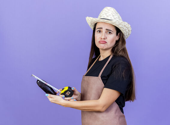 制服悲伤美丽的园丁女孩穿着制服戴着园艺帽用卷尺测量茄子隔离在蓝色背景上胶带帽子悲伤