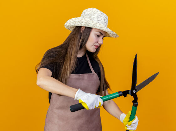 壁板看着一边想着美丽的园丁女孩穿着制服 戴着园艺帽 戴着手套 拿着剪子在一边孤立的橙色背景上思想女孩手套
