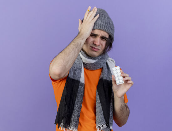 围巾戴着冬天帽子戴着围巾拿着药片手放在疼痛的头上隔离在紫色背景上的年轻病患头不愉快生病