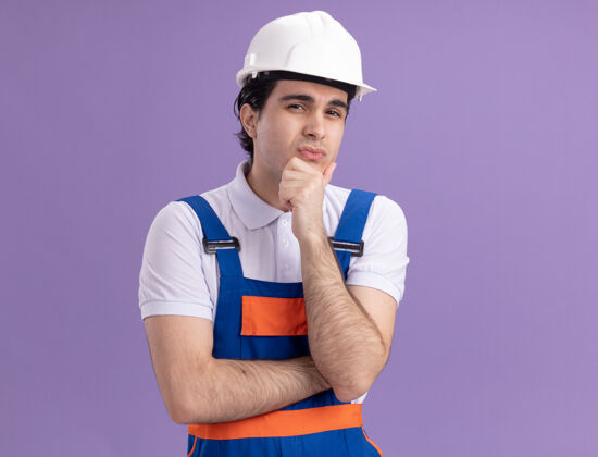 建设者年轻的建筑工人穿着建筑制服 戴着安全帽 手放在下巴上 站在紫色的墙上思考着前方下巴头盔站立