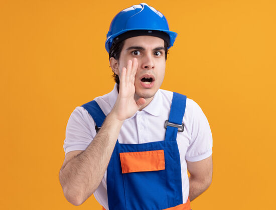 安全年轻的建筑工人穿着建筑制服 戴着安全帽 站在橙色的墙上 用手和嘴低声说着一个秘密制服男人头盔