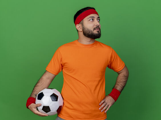 壁板看着侧面 这位戴着头带和腕带的年轻运动型男子在绿色背景下把球放在臀部年轻腕带球