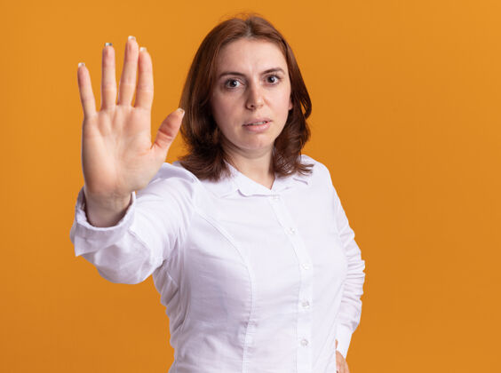 做身穿白衬衫的年轻女子严肃地看着前面 用手站在橘色的墙上做了个停车的手势手严肃女人