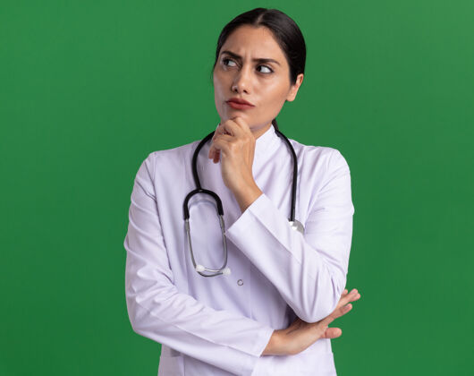 手年轻的女医生穿着医用外套 脖子上戴着听诊器 面带沉思的表情 手放在下巴上 站在绿色的墙上思考外套围着医生