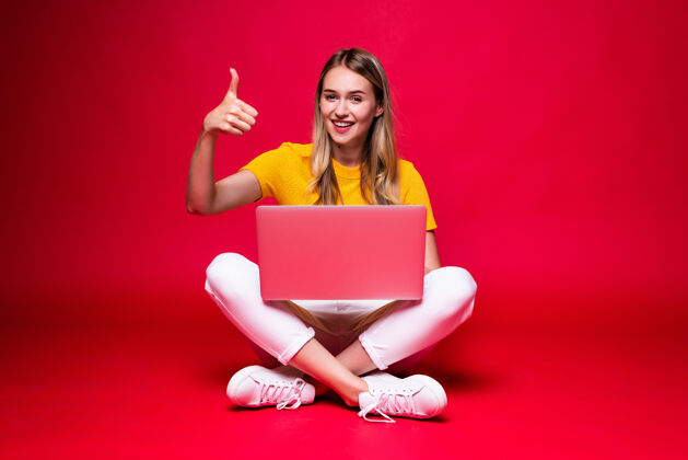 使用快乐的年轻卷发美女盘腿坐在地板上 在红墙上用笔记本电脑青少年笔记本电脑年轻