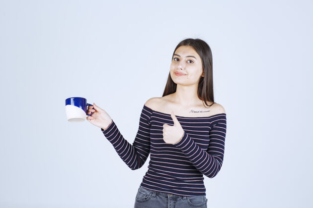 成功穿着条纹衬衫的女孩拿着一个咖啡杯 展示着享受的标志年轻休闲喜欢
