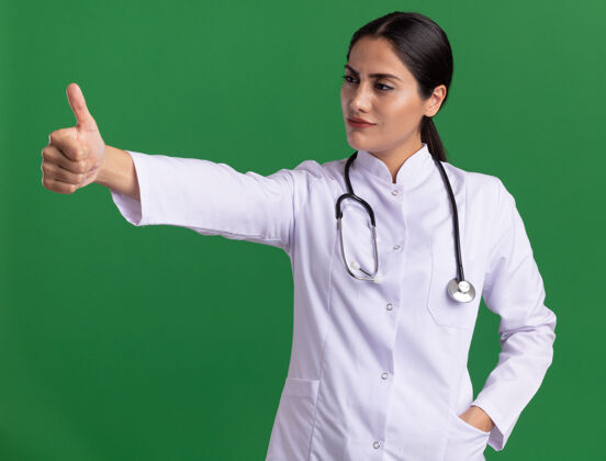 女士身穿医用外套 手持听诊器 表情严肃自信的年轻女医生站在绿色墙壁上竖起大拇指严肃站立听诊器