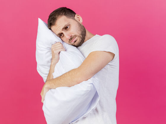 请看着身边高兴的年轻病夫抱着一个孤立在粉色背景上的枕头壁板疾病拥抱