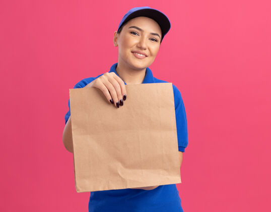 帽子快乐的年轻送货员 身穿蓝色制服 戴着帽子 手里拿着纸包 站在粉红色的墙上 面带微笑地看着前面欢呼制服纸