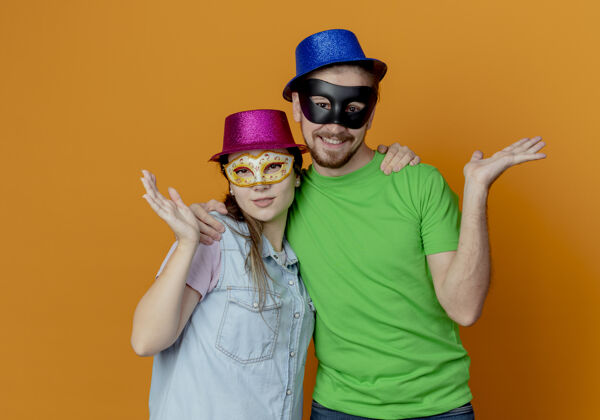 女人一对年轻的夫妇戴着粉色和蓝色的帽子戴上化装眼罩举起手看着橙色的墙上孤立手面具派对