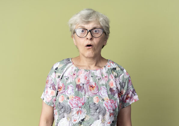 老人惊讶的戴眼镜的老妇人在橄榄绿的墙上显得孤立无援橄榄色老人眼镜