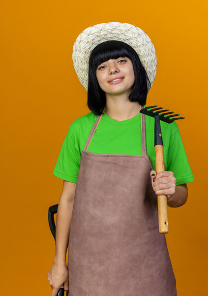 女性自信微笑的年轻女园丁穿着制服 戴着园艺帽 手里拿着铲子和耙子制服橙色耙