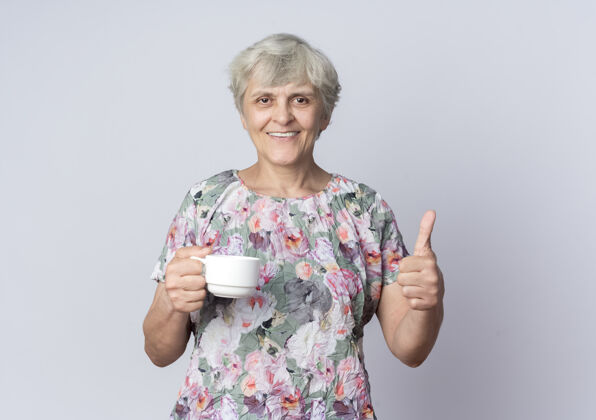 女人微笑的老妇人举着杯子 竖起大拇指孤立地站在白墙上老人拇指微笑