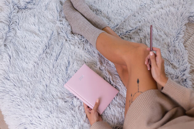 腿穿着羊毛袜子和毛衣的女人 粉色笔记本上写着2021 臀部有大纹身女人坐在卧室的家里的床上钢笔爱好住宿