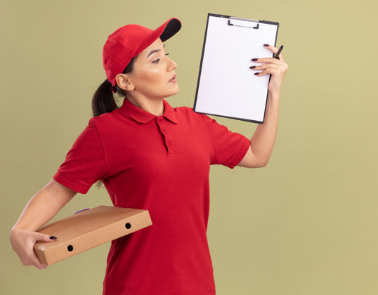 披萨身穿红色制服 头戴鸭舌帽的年轻送货员站在绿色的墙上 手里拿着披萨盒 上面展示着剪贴板和白纸 看起来很自信送货剪贴板女人