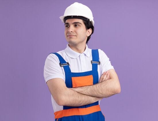 男人年轻的建筑工人身穿建筑制服 戴着安全帽 双手交叉 自信地看着一边 站在紫色的墙上手臂年轻安全