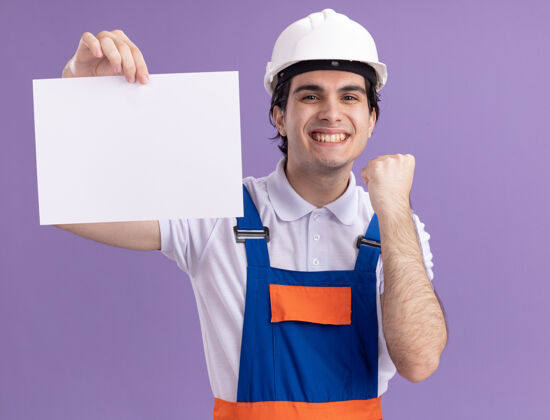 安全快乐的年轻建筑工人穿着建筑制服 戴着安全帽 拿着空白页 面带微笑地看着前面 握紧拳头站在紫色的墙上制服页面年轻