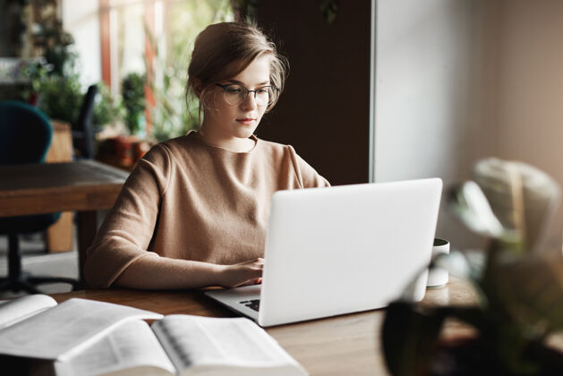现代工作 生活方式和商业理念戴着时髦眼镜的漂亮专注的欧洲女性坐在笔记本电脑旁的咖啡馆里 在笔记本上工作 围着书 做笔记企业家咖啡无线