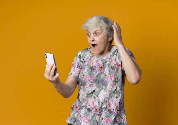 惊喜惊讶的老妇人举手看着隔离在橙色墙上的电话手起来提高