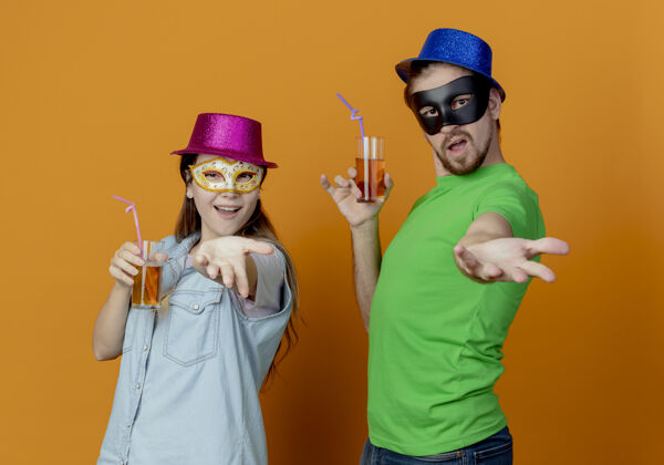 蓝色年轻快乐的夫妇戴着粉色和蓝色的帽子戴上化装眼罩拿着一杯果汁在橘色的墙上伸出孤立的手派对欢乐粉色