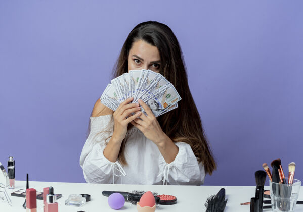 紫色惊讶的美丽女孩坐在桌子旁 拿着化妆工具 看着紫色墙上孤立的钱女人工具桌子
