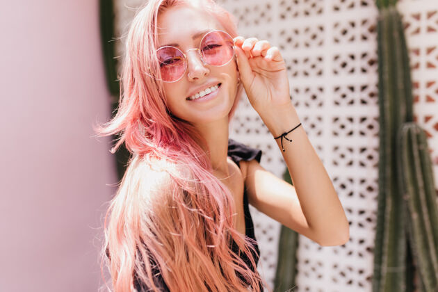 年轻高兴的皮肤黝黑的白种女人 粉红色的头发高兴的欧洲女士触摸她的太阳镜 而与异国植物摆姿势金发室内迷人