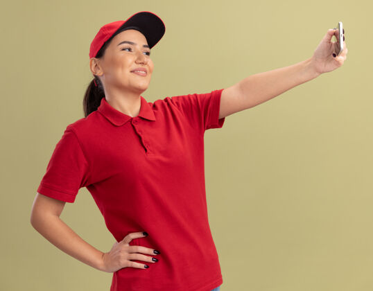 女人身穿红色制服 戴着帽子的年轻送货员正在用智能手机自拍 站在绿色的墙边 开心地微笑着帽子微笑制服