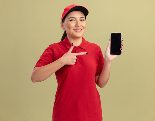 微笑年轻的送货员身穿红色制服 手持智能手机 用食指指着它微笑着自信地站在绿色的墙上制服帽子目录