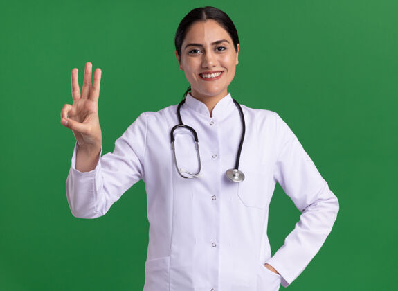 听诊器穿着医用外套的年轻女医生拿着听诊器看着前面 指着绿色的墙上站着的三号手指医生三个手指