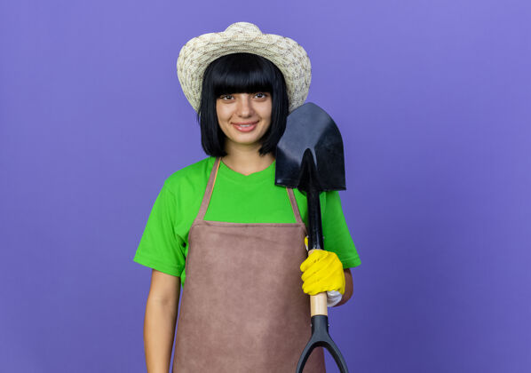 黑桃微笑的年轻女园丁穿着制服戴着园艺帽和手套手持铁锹园艺花园年轻