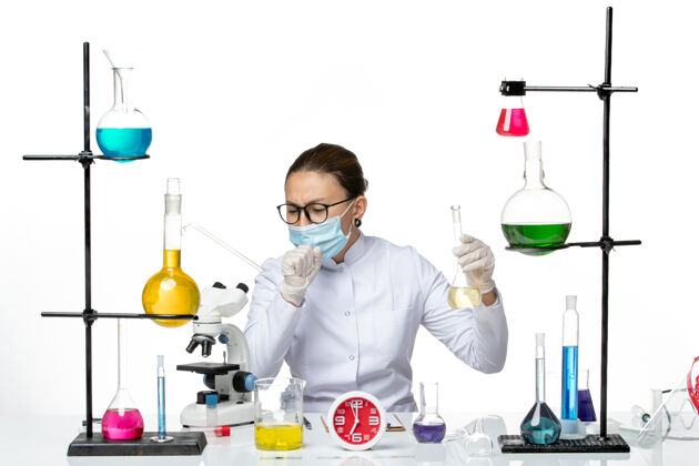 溶液前视图穿着白色医疗服的女化学家戴着口罩拿着药水在白色背景上咳嗽化学家实验室病毒冠状病毒-飞溅药物前面面罩