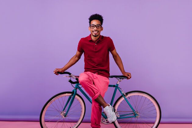 成人穿着粉色裤子坐在自行车上的快乐黑人男模戴眼镜的非洲男孩的室内笑镜头时尚兴奋活动