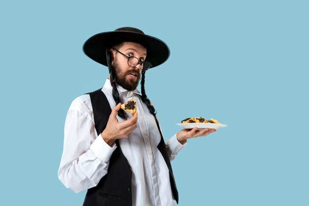 律法这位年轻的东正教犹太男子戴着黑帽子 在犹太普瑞姆节上吃哈曼塔森饼干耳朵甜点盘子