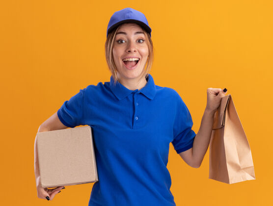 年轻兴奋的年轻漂亮的送货女孩在制服举行纸包装和名片盒在橙色制服漂亮摆姿势