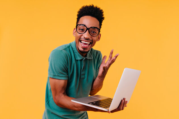 黄色帅气的戴眼镜的男自由职业者微笑着欣喜若狂的非洲学生拿着笔记本电脑 表达着幸福积极模型美国