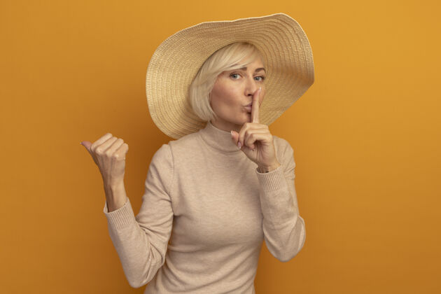 漂亮自信的金发斯拉夫女人 戴着沙滩帽 做着沉默的手势 指着橙色的边自信时尚斯拉夫
