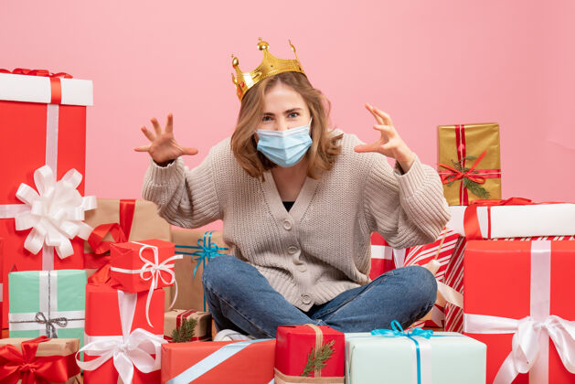 圣诞节正面图：坐在圣诞节周围的年轻女性戴着无菌面具出现面具粉色礼物